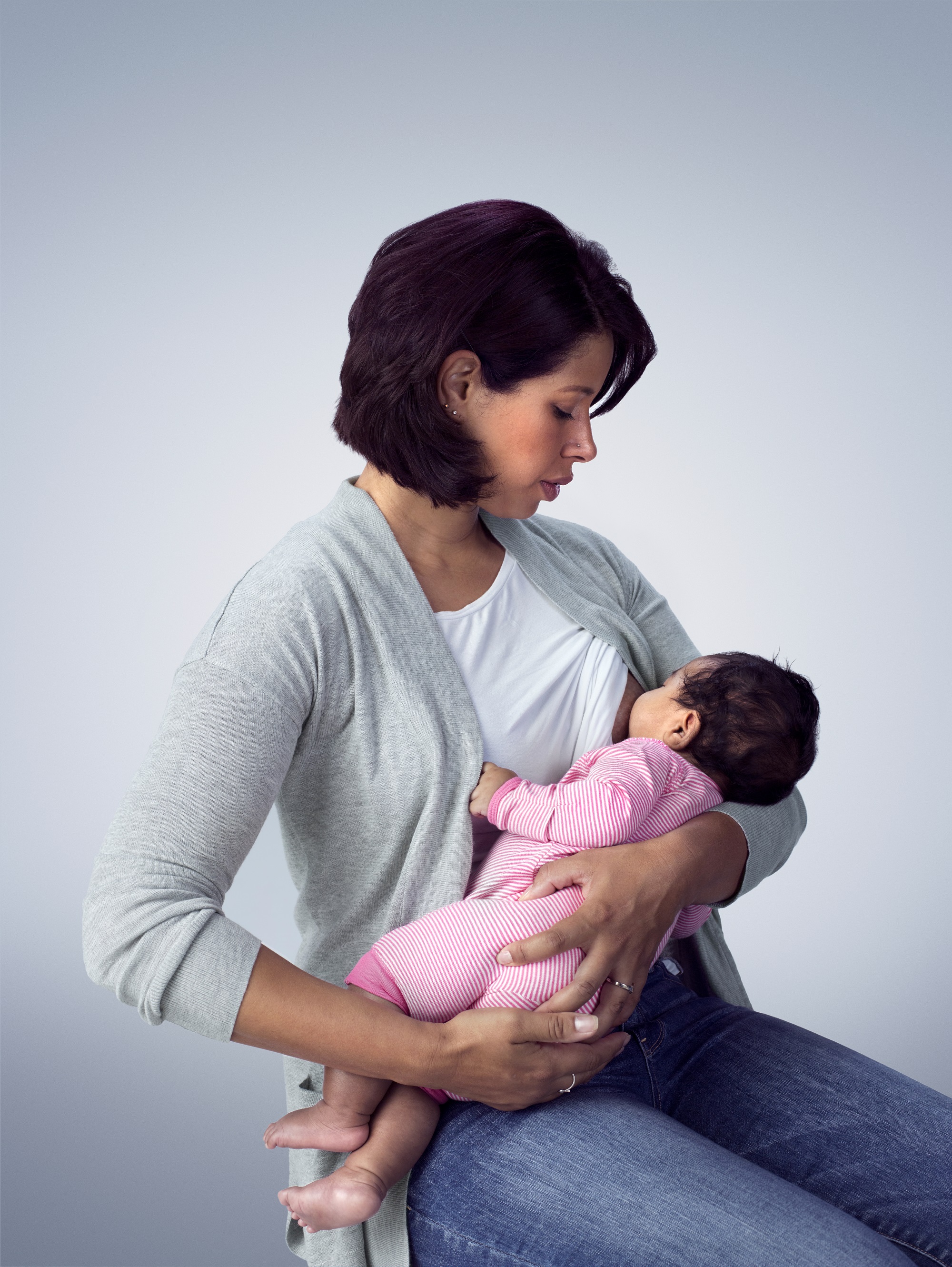 A woman in a gray cardigan is nursing a baby in a pink outfit, both looking content.