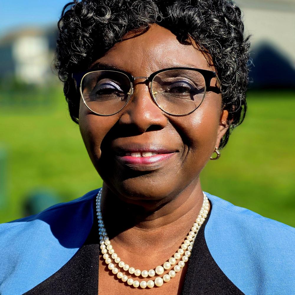 A woman with short curly hair, glasses, and a pearl necklace smiles in a sunny outdoor setting.