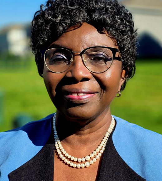 A woman with short curly hair, glasses, and a pearl necklace smiles in a sunny outdoor setting.