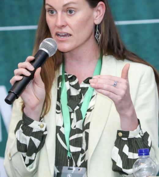 Woman speaking into microphone at conference, green backdrop, animated gesture