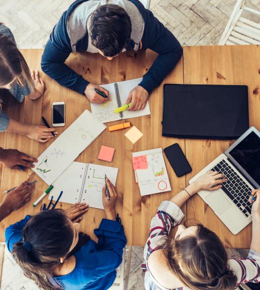 Overhead view of five people brainstorming around a wooden table with laptops and sticky notes