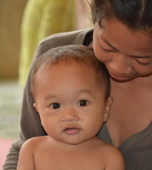 A woman lovingly holds a baby, both smiling softly, in a warm interior setting.
