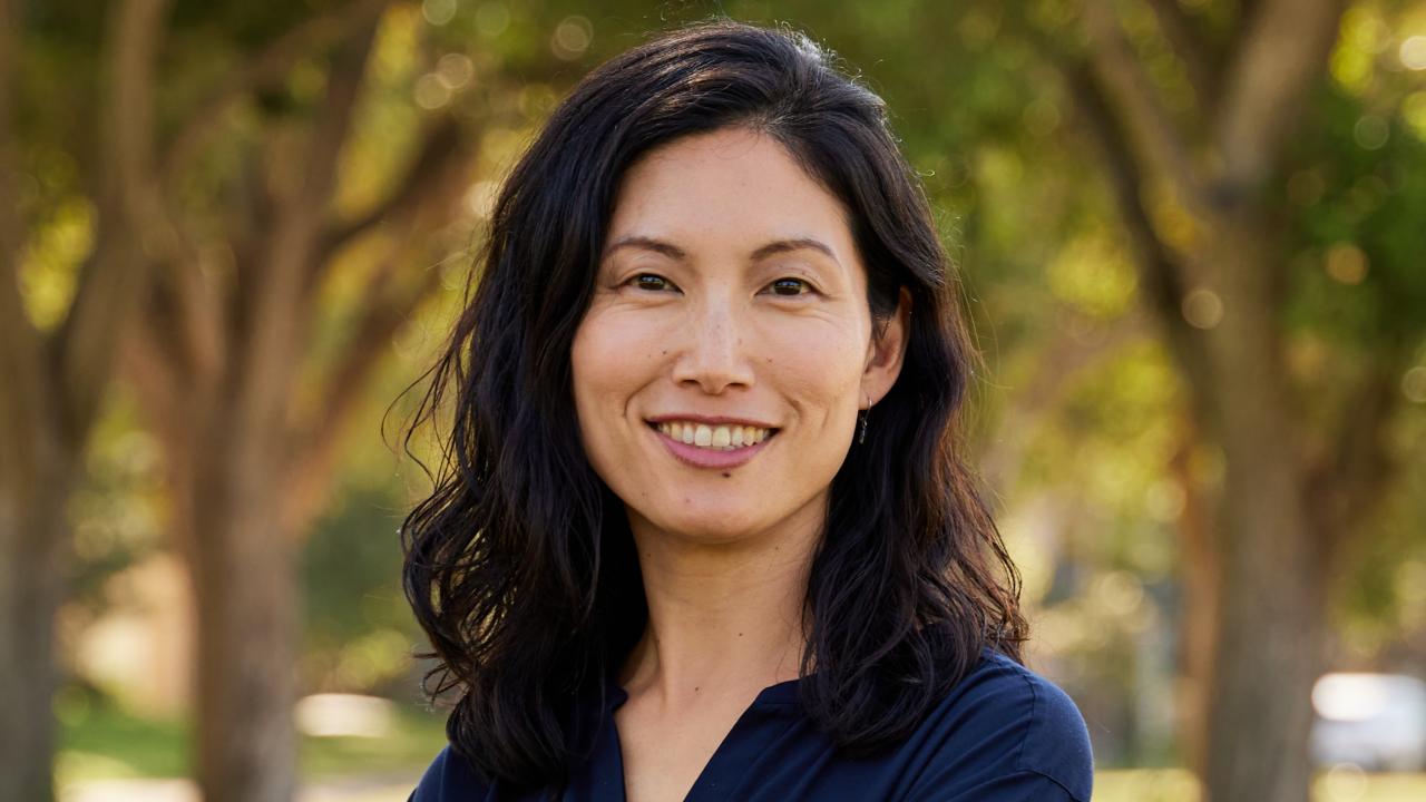 Katherine Kwon smiling, standing with arms folded on grass with trees in background
