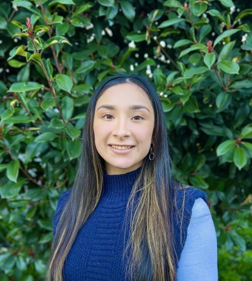 Andrea smiling in dark blue sweater and light blue long sleeves, standing in front of green high bushes