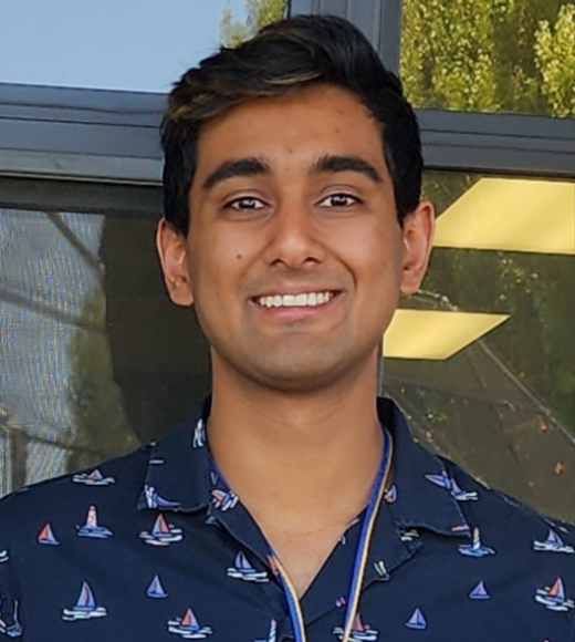 Hussain smiling, standing against windows in dark blue shirt with tiny sailboats