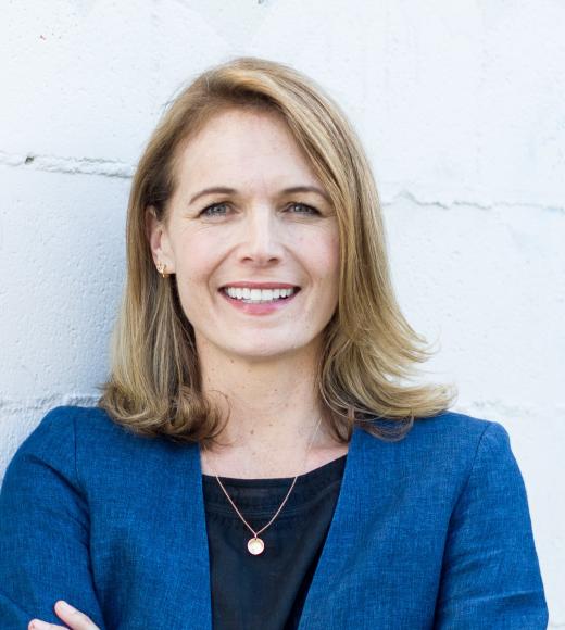 Lia in dark blue suit jacket, against gray white wall, smiling