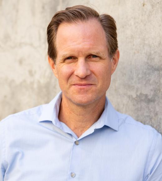 Travis leaning against cement wall, wearing light blue shirt, gazing at the camera