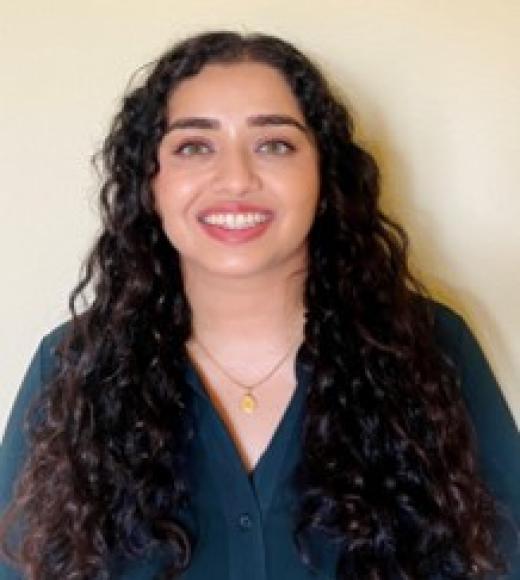 Hareem smiling against a white wall, in a black v-neck sweater, with long black hair
