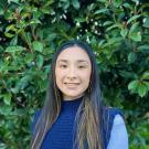 Andrea smiling in dark blue sweater and light blue long sleeves, standing in front of green high bushes