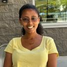 Lidya Abebe sitting outside in yellow shirt & glasses, smiling.