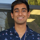 Hussain smiling, standing against windows in dark blue shirt with tiny sailboats