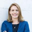 Lia in dark blue suit jacket, against gray white wall, smiling