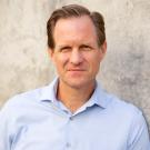 Travis leaning against cement wall, wearing light blue shirt, gazing at the camera