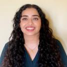 Hareem smiling against a white wall, in a black v-neck sweater, with long black hair